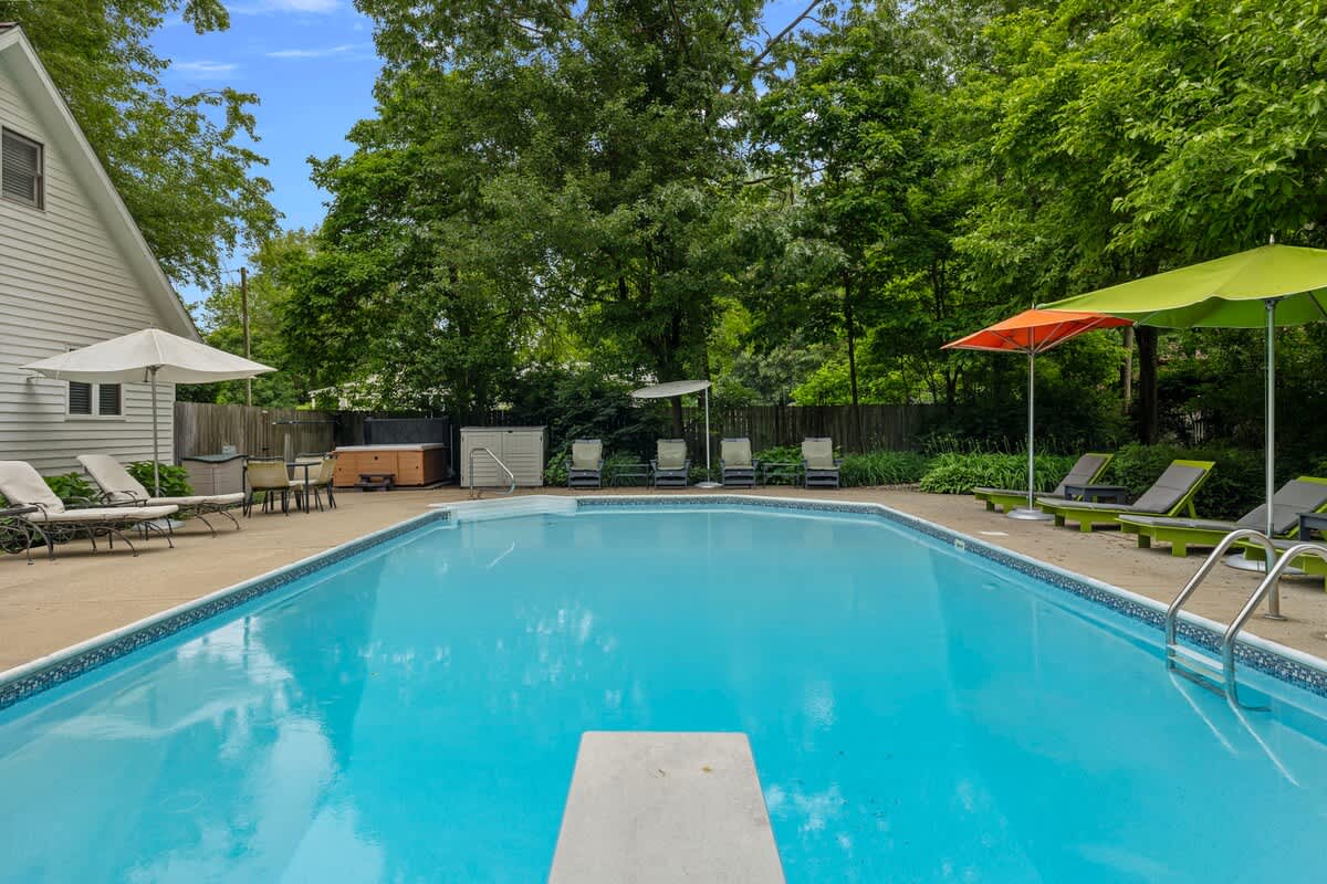 An inviting blue swimming pool with a diving board in the foreground, surrounded by a concrete deck with lounge chairs, umbrellas, and a hot tub visible in the background, all nestled among dense green trees.