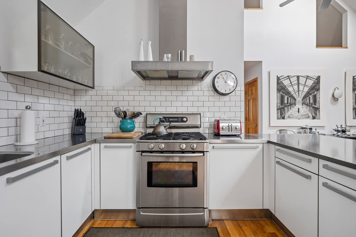 Close-up view of a modern kitchen with white flat-panel cabinets, stainless steel countertops, a gas range, and a subway tile backsplash, accented by a red toaster and a blue kettle.