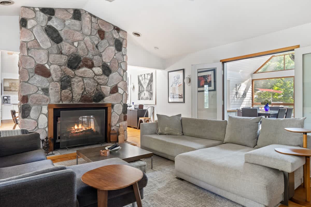 The living room showcases a large stone fireplace and two light-colored sectional sofas arranged around a dark coffee table. Sliding glass doors open to a bright screened porch, connecting indoor and outdoor living spaces.