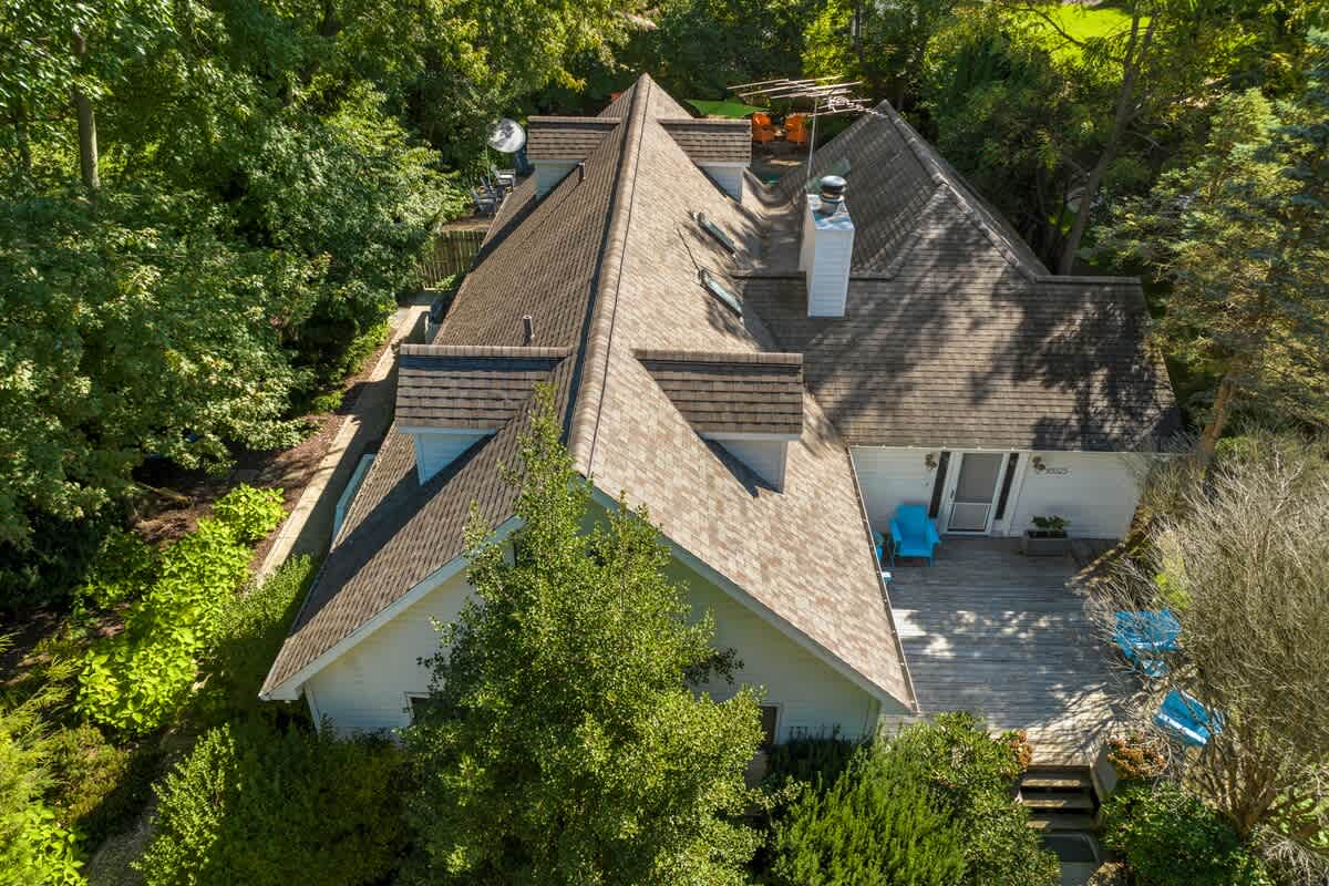 An aerial perspective highlighting the complex brown gabled roof of the house, surrounded by abundant green trees, with a wooden deck and outdoor furniture visible below.