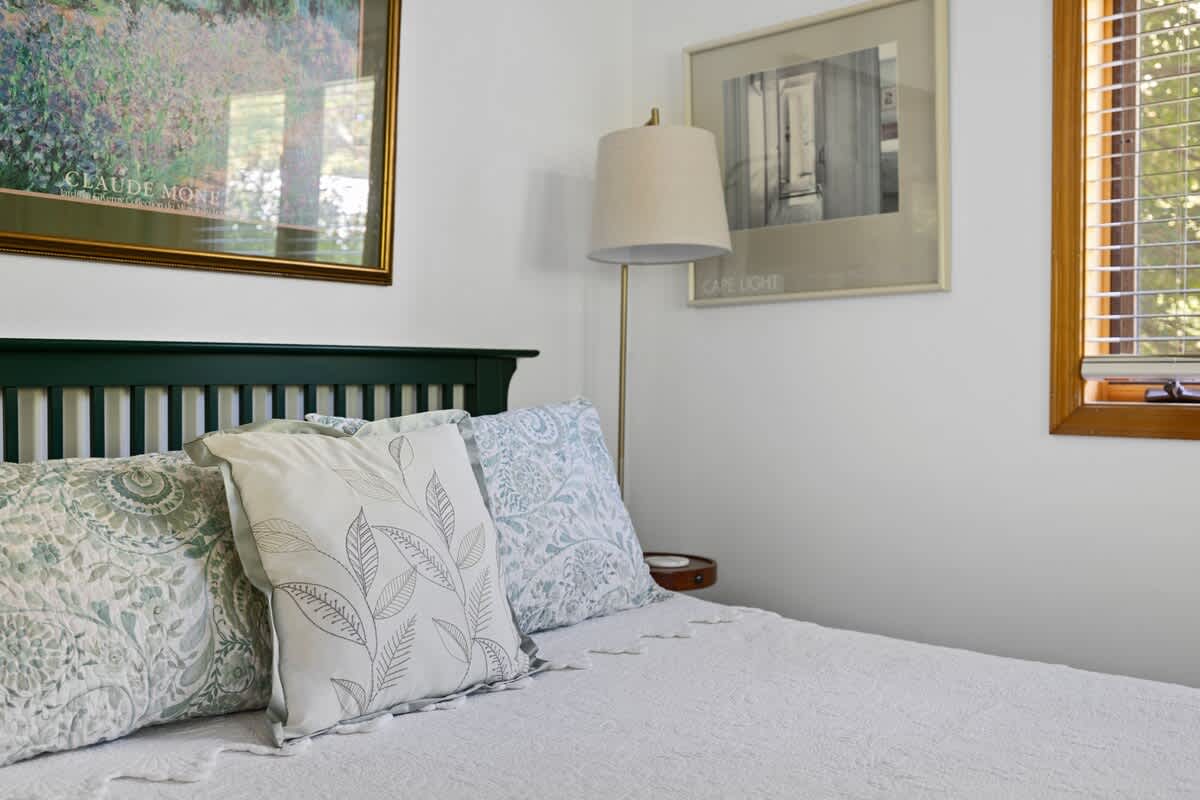 Close-up of a queen bed with a dark green wooden headboard, white quilted bedding, and light blue and white patterned throw pillows. A green nightstand with a lamp and framed art prints are visible on the white wall.