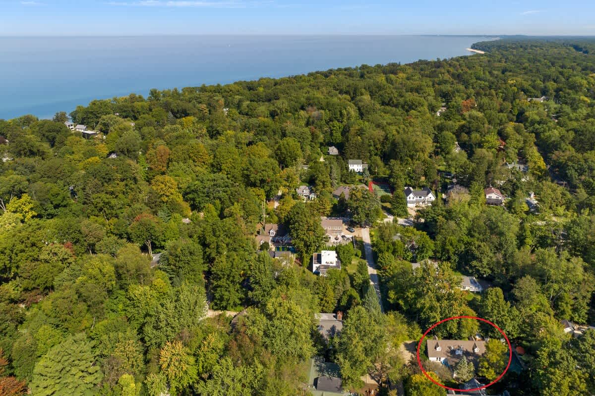 A wide aerial shot displaying the property, circled in red, nestled within a dense forest of green trees, with a large body of water (likely a lake or ocean) visible in the distant background under a clear sky.