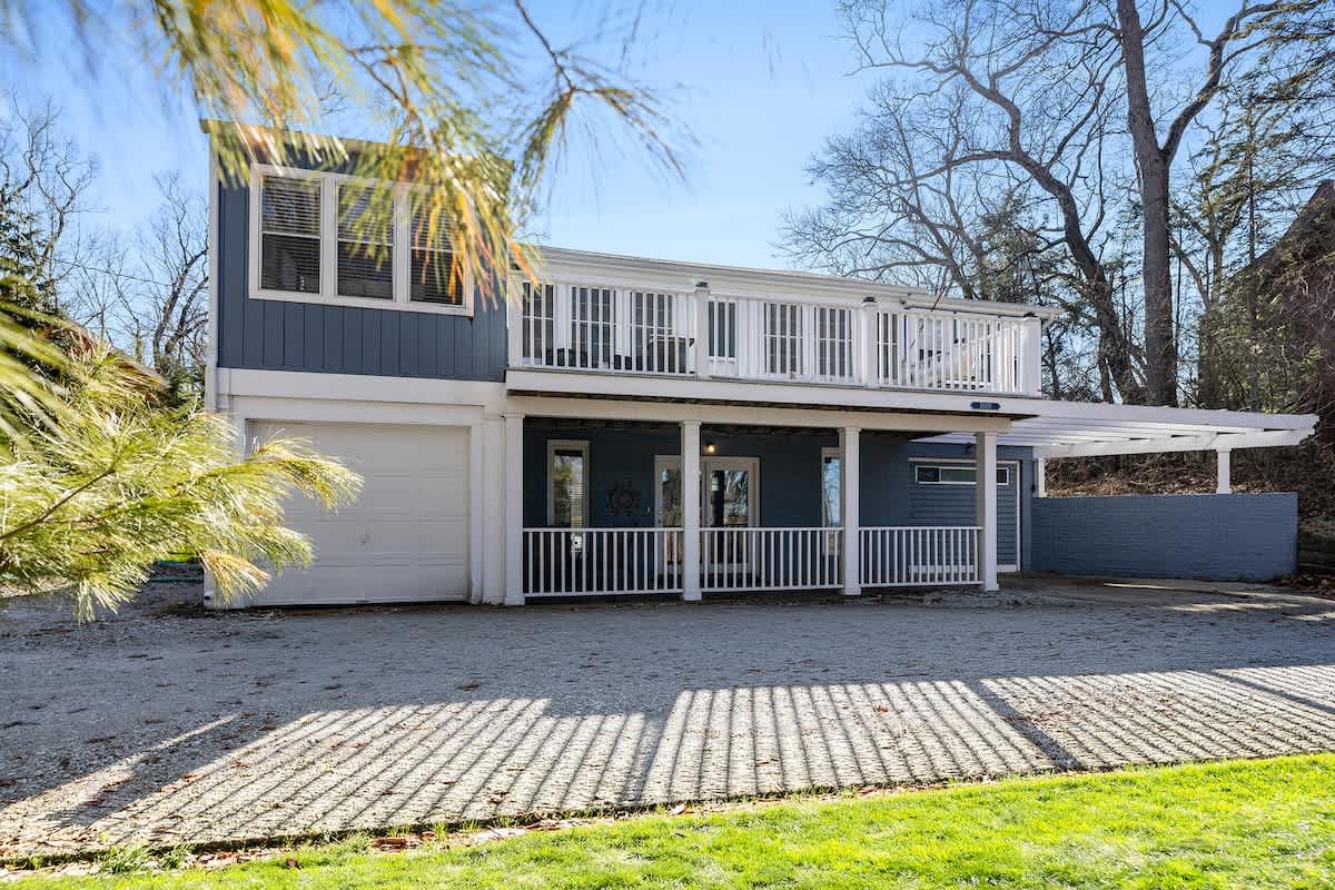 Front exterior of a two-story blue house with white trim and a second-story balcony with white railings, featuring a covered porch on the first floor and a gravel driveway in front.