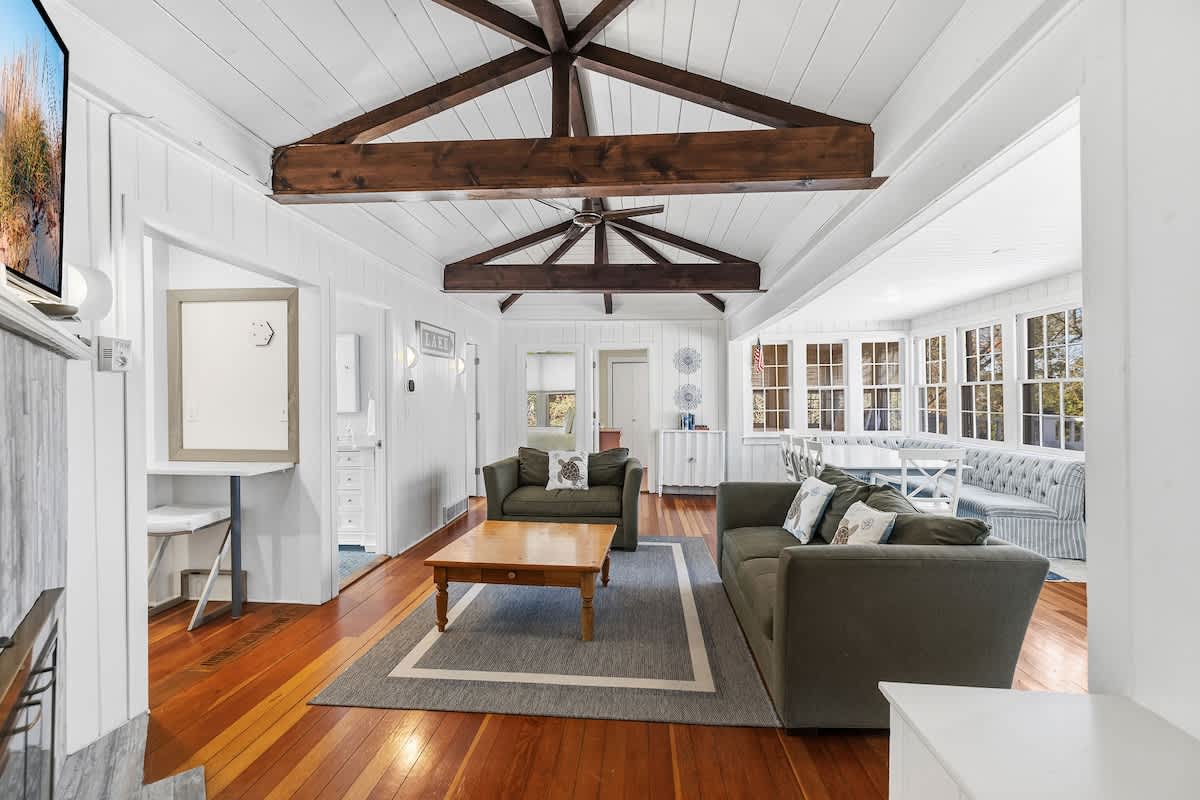 Bright living room with two olive green sofas, a wooden coffee table on a grey rug, and a dining area with a built-in bench seating visible through an archway. The room features a white plank ceiling with dark wood exposed beams and hardwood floors.