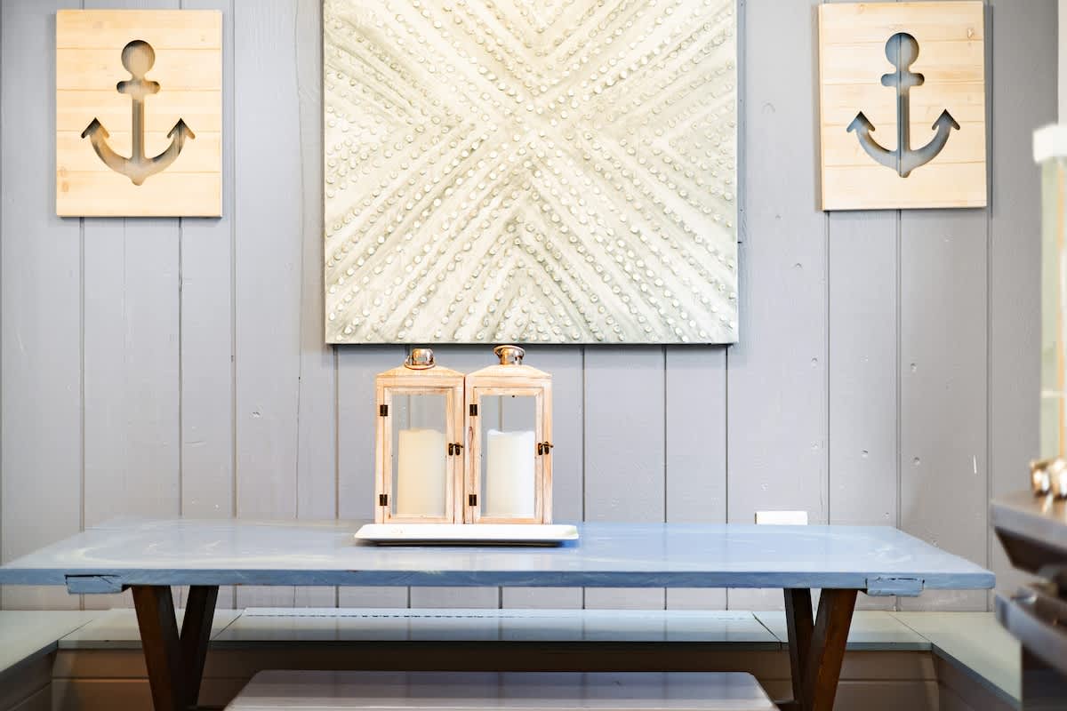 A light gray shiplap wall adorned with two wooden anchor decorations and a textured square artwork, above a light blue console table with two decorative lanterns, creating a nautical-themed accent space.