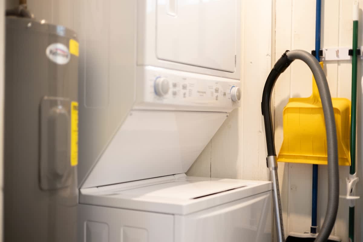 Compact laundry area featuring a white stacked washer and dryer unit next to a water heater. Cleaning supplies, including a vacuum hose and dustpan, are neatly hung on the wall.