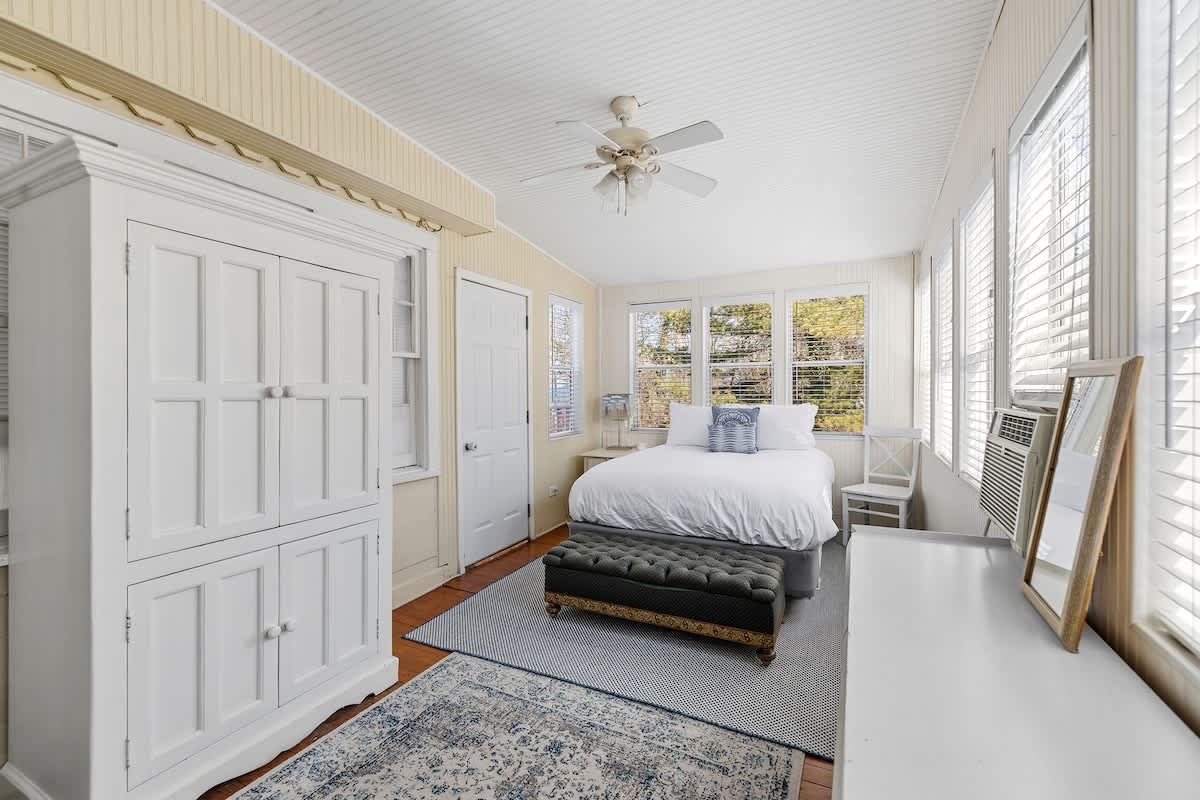 A spacious bedroom with light-colored paneled walls, a white bed, a green tufted bench, a large white armoire, and multiple windows with blinds, all under a white ceiling fan.