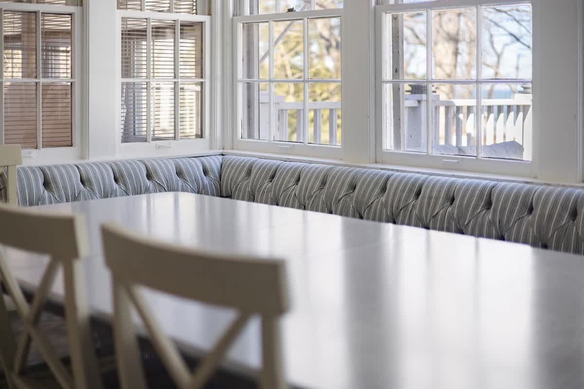 Bright dining area featuring a long white dining table with white cross-back chairs and a striped built-in banquette seating along a wall of large windows. The windows offer views of the outdoors.