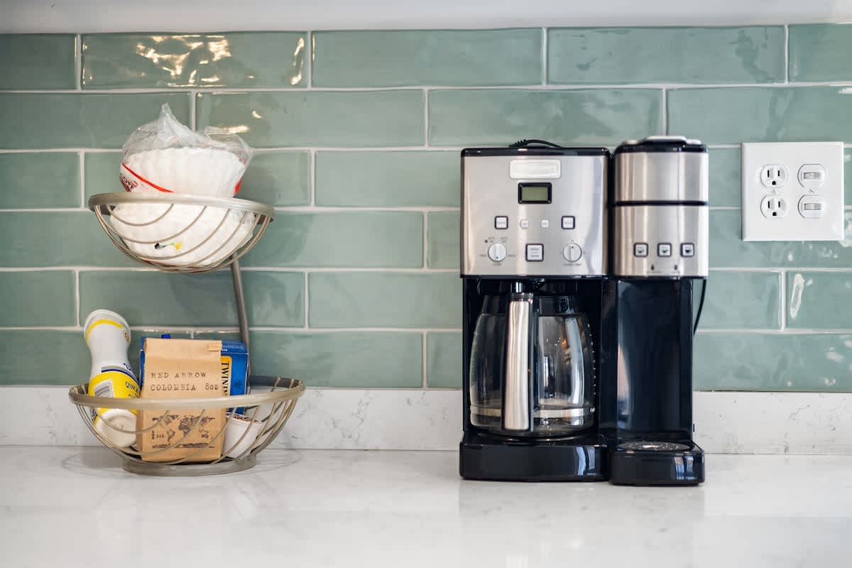 Close-up of a kitchen counter with a coffee maker, toaster, and a two-tiered fruit basket filled with coffee filters and coffee. The light green subway tile backsplash and white countertop are visible.