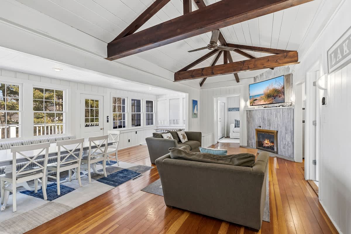 Bright open-concept living and dining area with white shiplap walls, vaulted ceilings with dark wood beams, hardwood floors, a dining table with white chairs, and a living space with two gray sofas facing a wall-mounted TV above a fireplace.