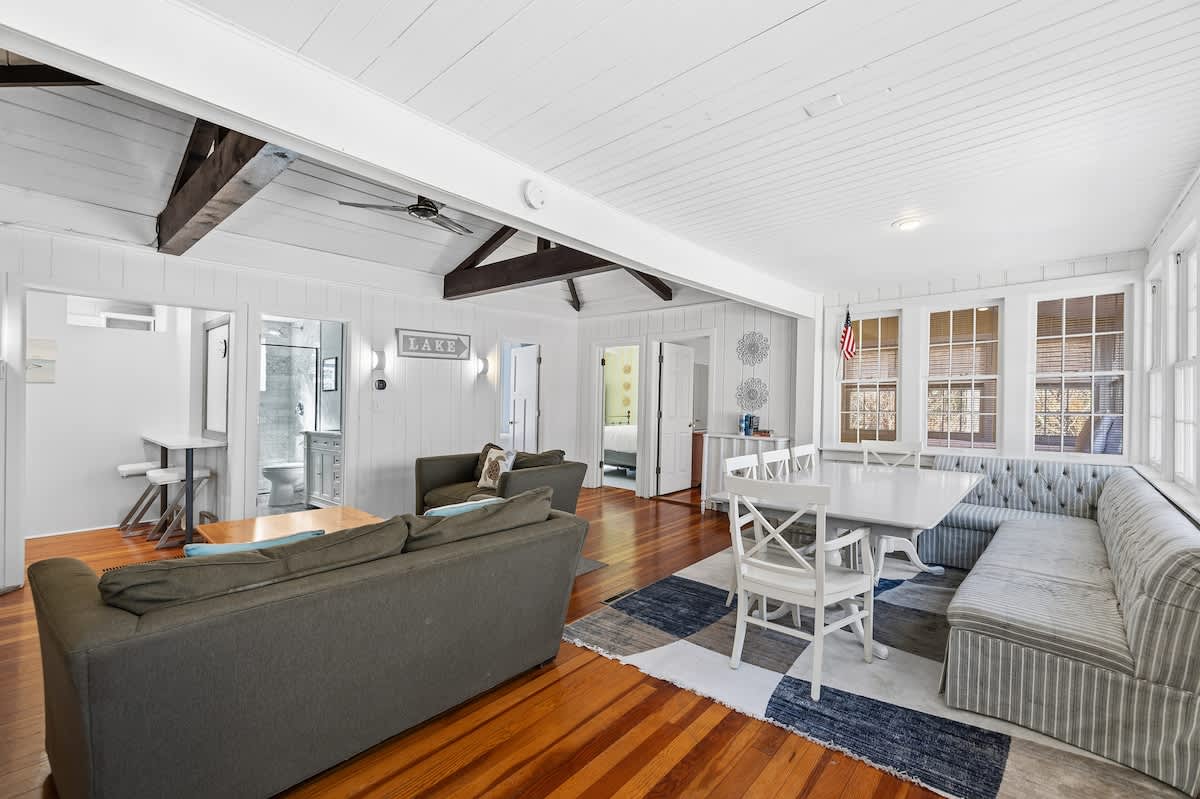 Wide shot of the open-concept living and dining area, featuring two olive green sofas, a wooden coffee table, and a dining nook with a striped built-in bench and white chairs. The room has hardwood floors, white plank walls, and a white plank ceiling with dark wood exposed beams.