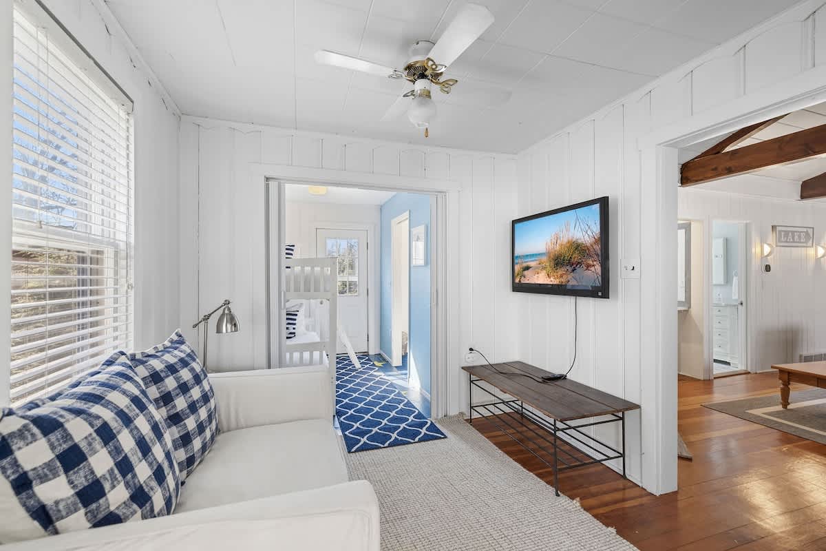 A bright living area with white paneled walls, a white sofa with blue plaid pillows, a wall-mounted TV, and an open doorway leading to a children's room with bunk beds.