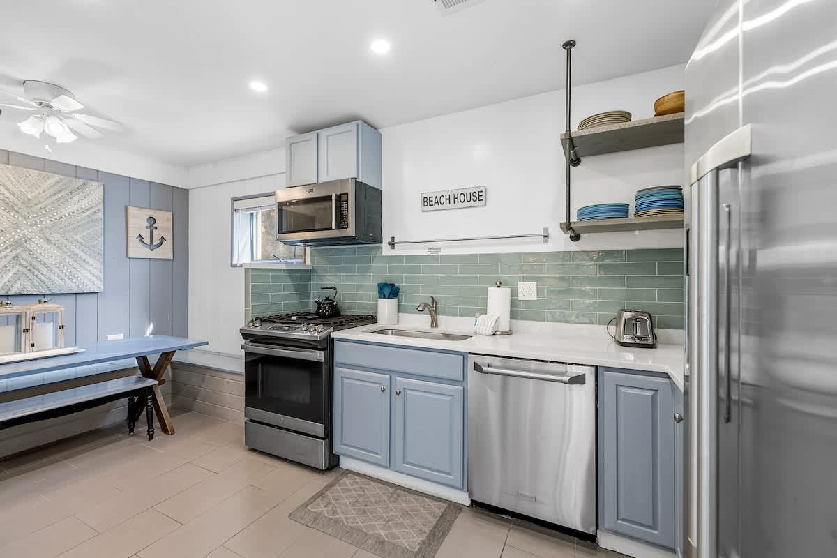 Kitchen with light blue cabinets, white countertops, and a light green subway tile backsplash, featuring a stainless steel gas range, microwave, and dishwasher. A small dining area with a blue picnic-style table and bench seating is visible to the left.