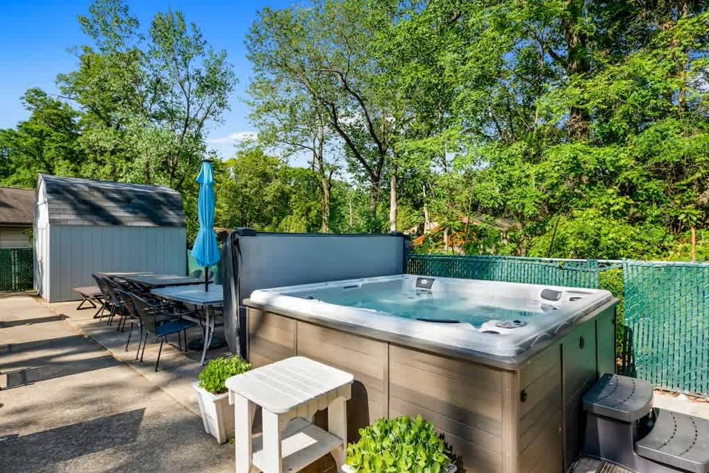 Outdoor patio area featuring a large bubbling hot tub with a dark brown exterior, an outdoor dining table with chairs, and a small shed in the background, all surrounded by lush greenery.