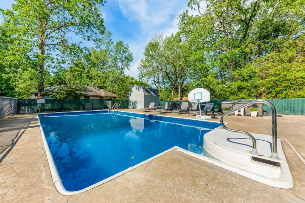 Large rectangular inground pool with a bright blue liner, surrounded by a concrete deck with lounge chairs and a basketball hoop, enclosed by a green chain-link fence and mature trees.
