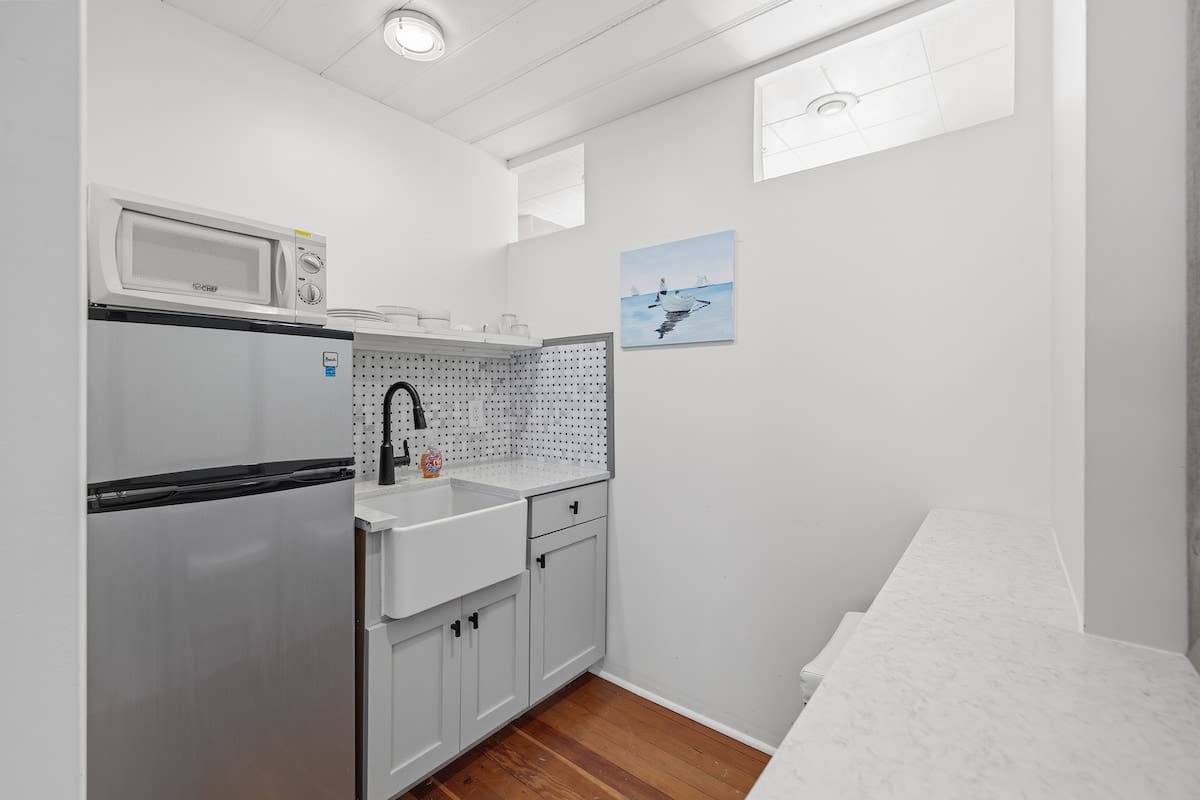 Compact kitchenette featuring a mini-fridge, microwave, farmhouse sink, and light grey cabinets with open shelving. The room has hardwood floors and white walls with a small window above the sink.