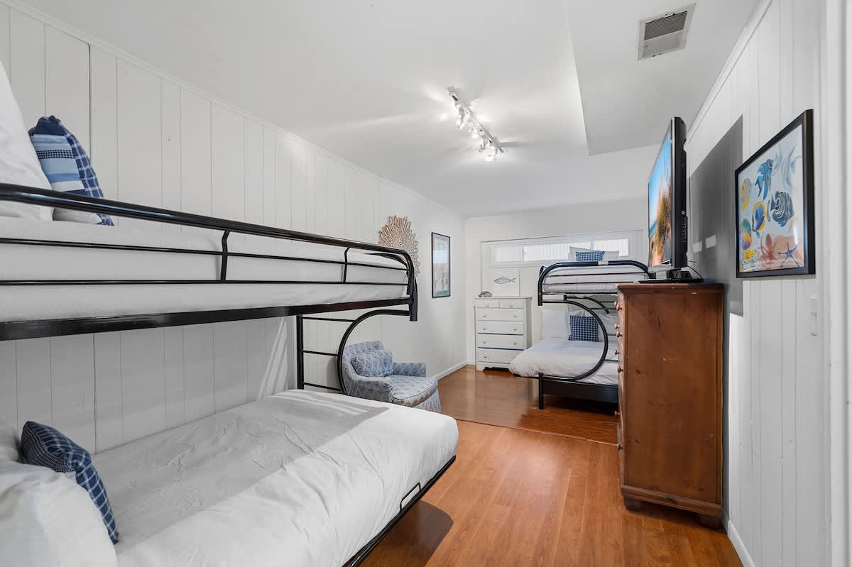 Spacious bedroom with light wood flooring and white paneled walls, featuring two black metal bunk beds with white bedding and blue plaid pillows. A large flat-screen TV is mounted on a dresser, and nautical-themed artwork adorns the walls.