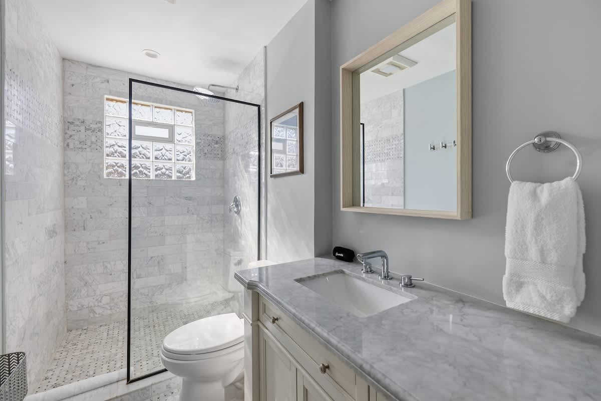 A modern bathroom with light gray walls, a walk-in shower featuring marble-patterned tiles and a glass enclosure, a white toilet, and a vanity with a gray marble countertop and single sink.