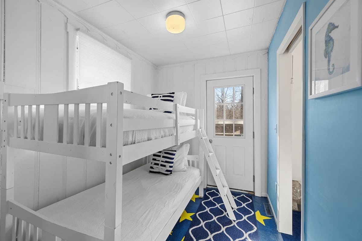 Bright white bedroom featuring a wooden bunk bed with white bedding and navy striped pillows, a blue and yellow star-patterned rug, and a white door leading outside. A seahorse artwork hangs on the blue accent wall.