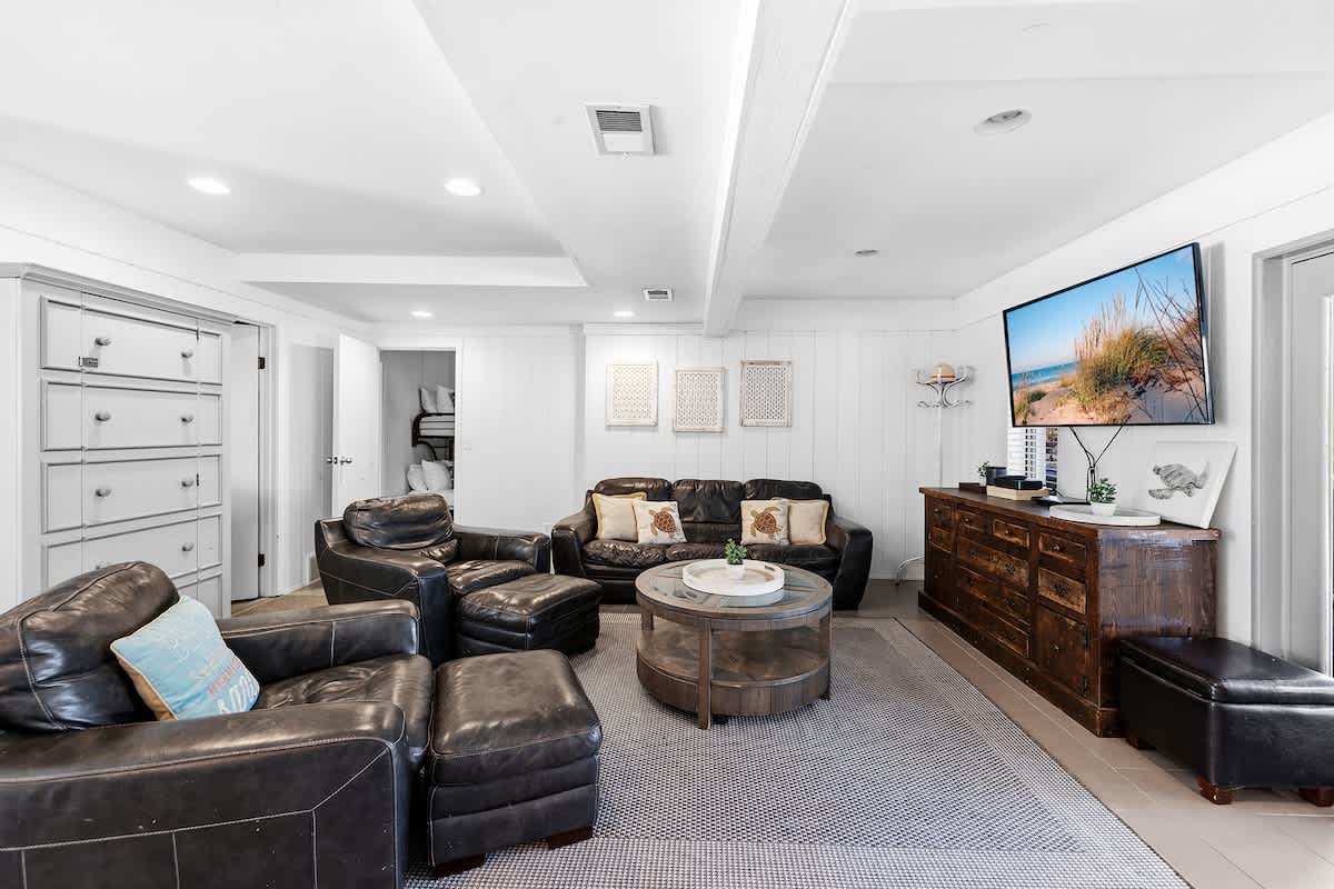 Cozy lower-level living room with dark leather seating, a round wooden coffee table, and a large wall-mounted TV above a rustic wooden console. The room has light tile flooring, white paneled walls, and a grey area rug.