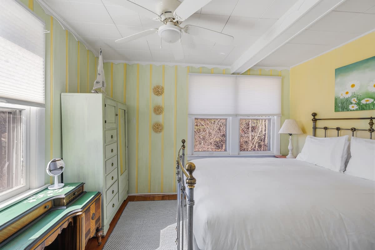 A bright bedroom featuring yellow and green striped walls, a brass bed, a light green dresser, a wooden desk, and multiple windows with white blinds, all under a white ceiling fan.