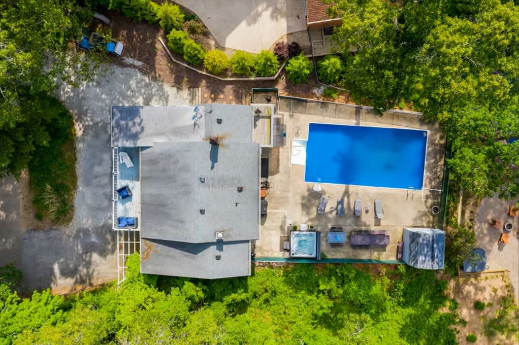 Aerial view of a house with a gray roof, surrounded by lush green trees. A large rectangular swimming pool and a hot tub are visible on a paved patio area next to the house.