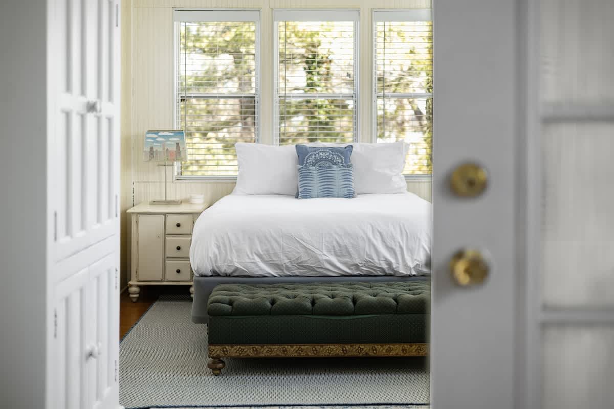 A bright bedroom featuring a white bed with a blue decorative pillow, a green tufted bench at the foot, and light-colored paneled walls with multiple windows providing natural light through blinds.