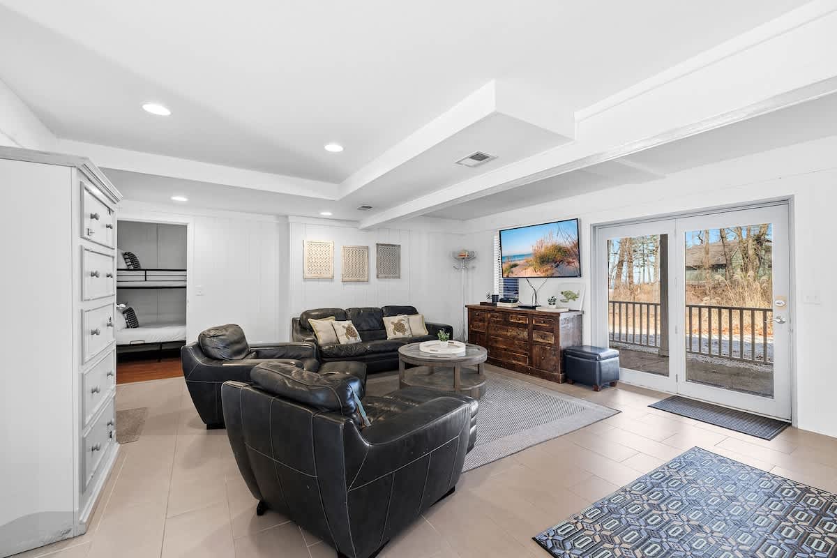 Spacious lower-level living room with dark leather sofas and armchairs, a round wooden coffee table, and a large wall-mounted TV. The room features light tile flooring, white paneled walls, and a door leading to an outdoor deck. Bunk beds are visible in an adjacent room.