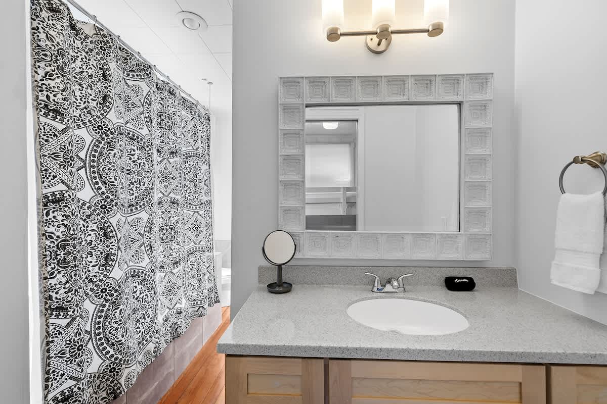 Modern bathroom with a light gray vanity, white oval sink, and a large mirror framed with decorative white tiles. A black and white patterned shower curtain is visible to the left, and a three-light vanity fixture illuminates the space.