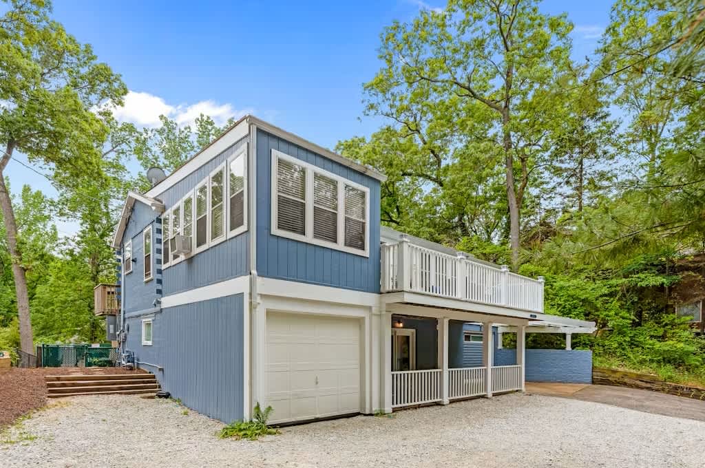 Two-story blue house with white trim and multiple windows, nestled among tall green trees. A white railing deck extends from the upper level, and a gravel driveway leads to a garage and carport.