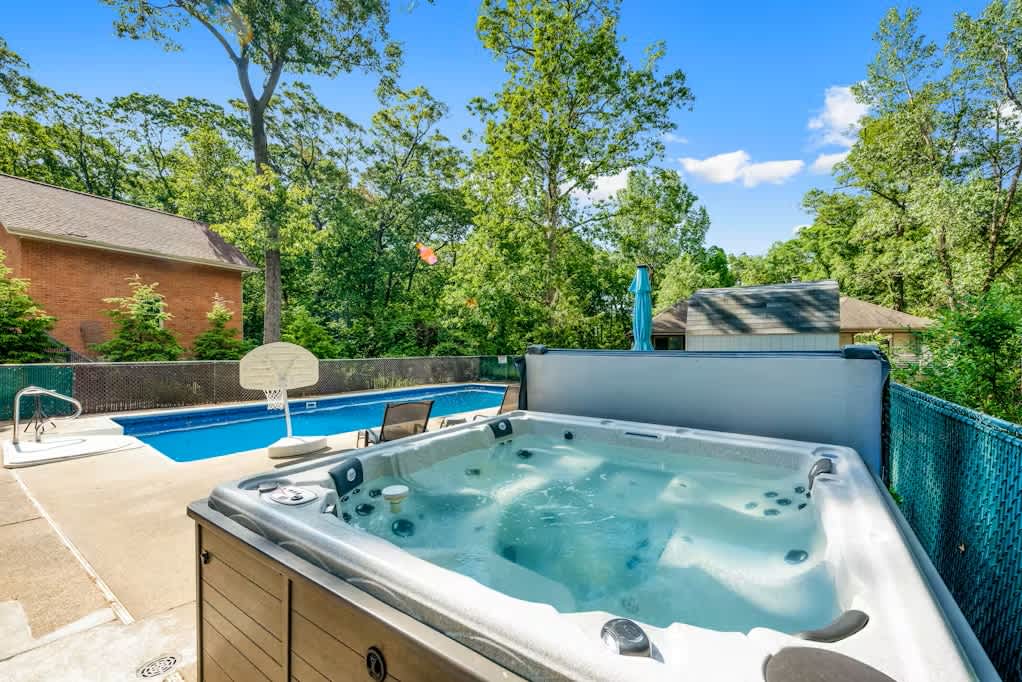 Outdoor patio area featuring a bubbling hot tub with a dark brown exterior, a blue inground pool with a basketball hoop, and a dining table with chairs, all surrounded by a chain-link fence and lush trees.