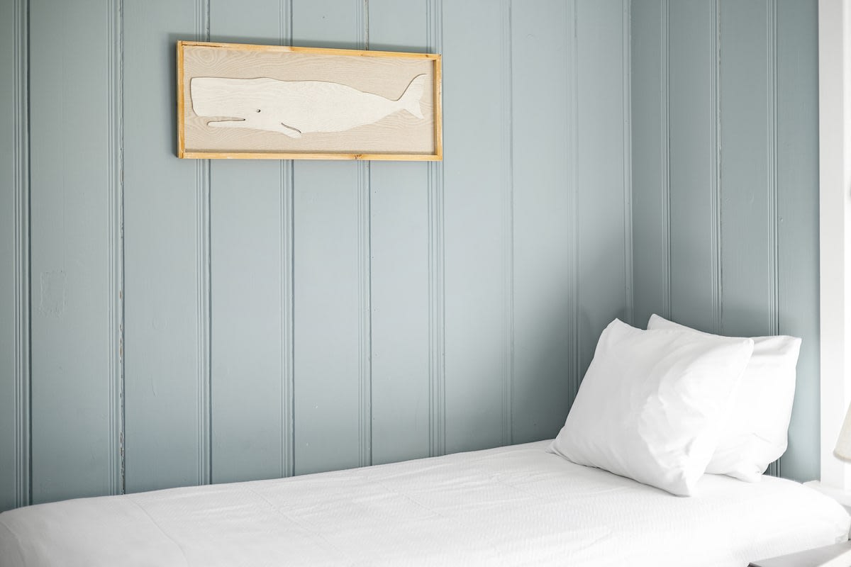A close-up view of one of the twin beds in a bedroom with light blue paneled walls, featuring white bedding and a framed whale artwork on the wall.