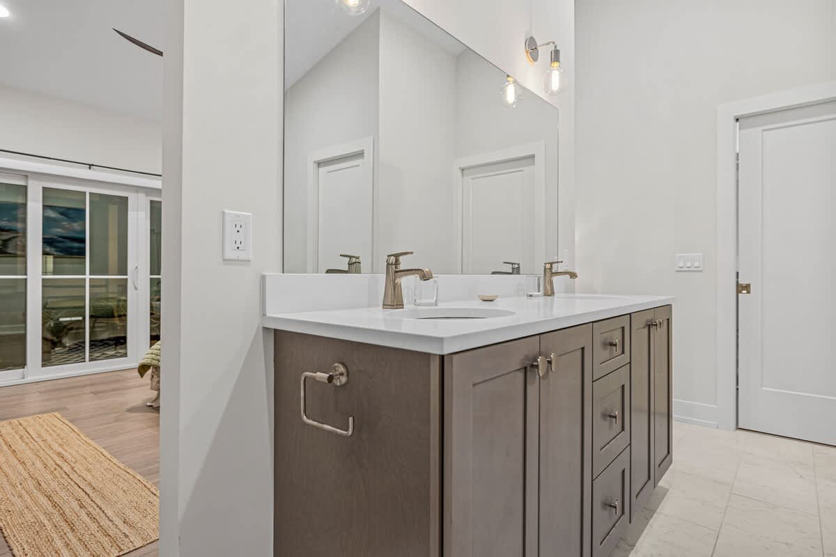 Modern bathroom featuring a double vanity with light gray cabinets, white countertops, and brushed gold fixtures. A large mirror extends across the wall, reflecting the adjacent bedroom.