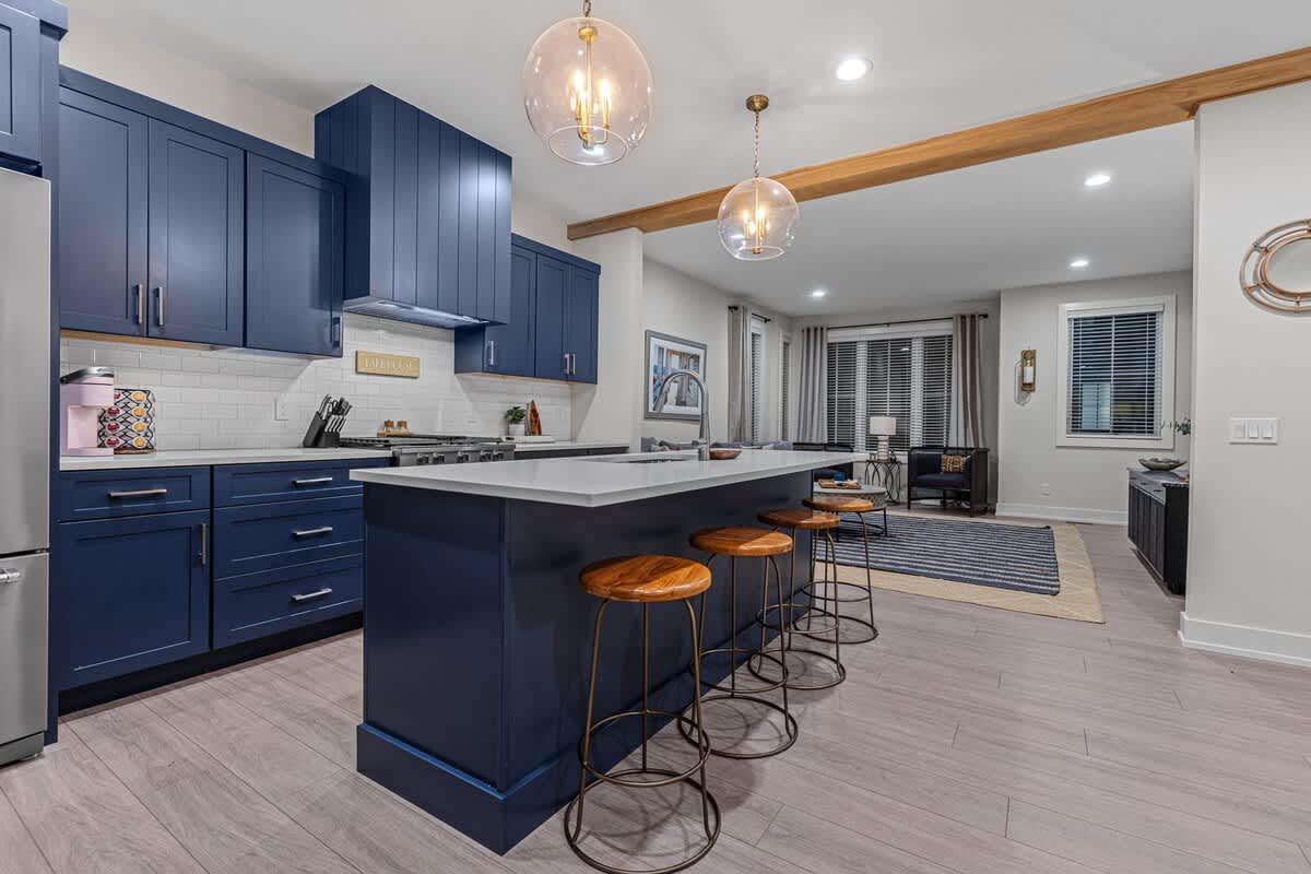 A modern kitchen with navy blue shaker-style cabinets, white subway tile backsplash, and light countertops, featuring a large island with a sink and four wooden bar stools. The living room is visible in the background through an open archway.