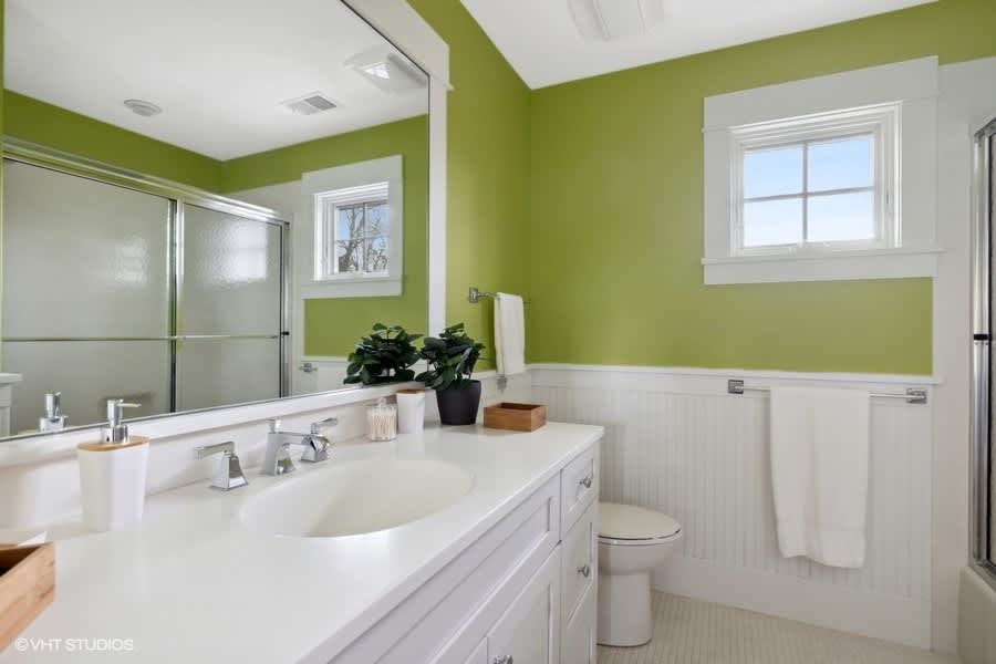 Bright bathroom with vibrant green walls and white wainscoting, featuring a double vanity with a white countertop, chrome fixtures, and a shower with a glass enclosure.