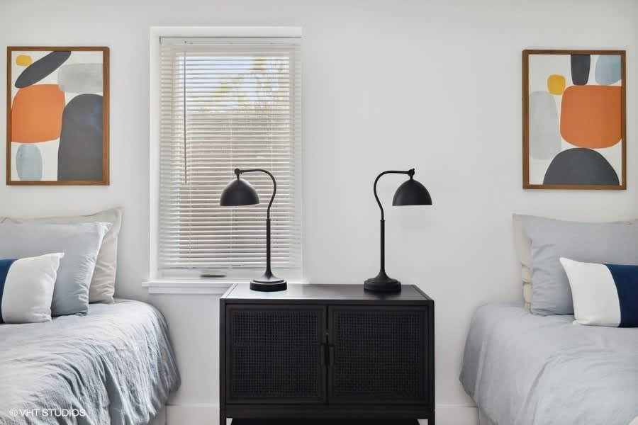 Close-up view of the space between two twin beds, featuring a black rattan-front cabinet with two black curved lamps, and a window with white blinds.