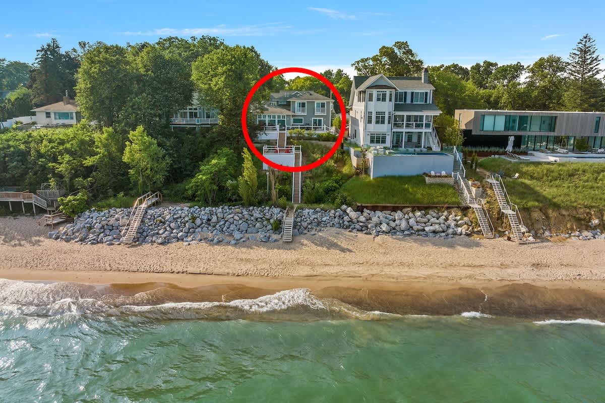 Aerial view of a gray multi-story house with a red circle highlighting it, situated on a green bluff overlooking a sandy beach and a large lake. Stairs lead down to the beach, flanked by other modern homes.