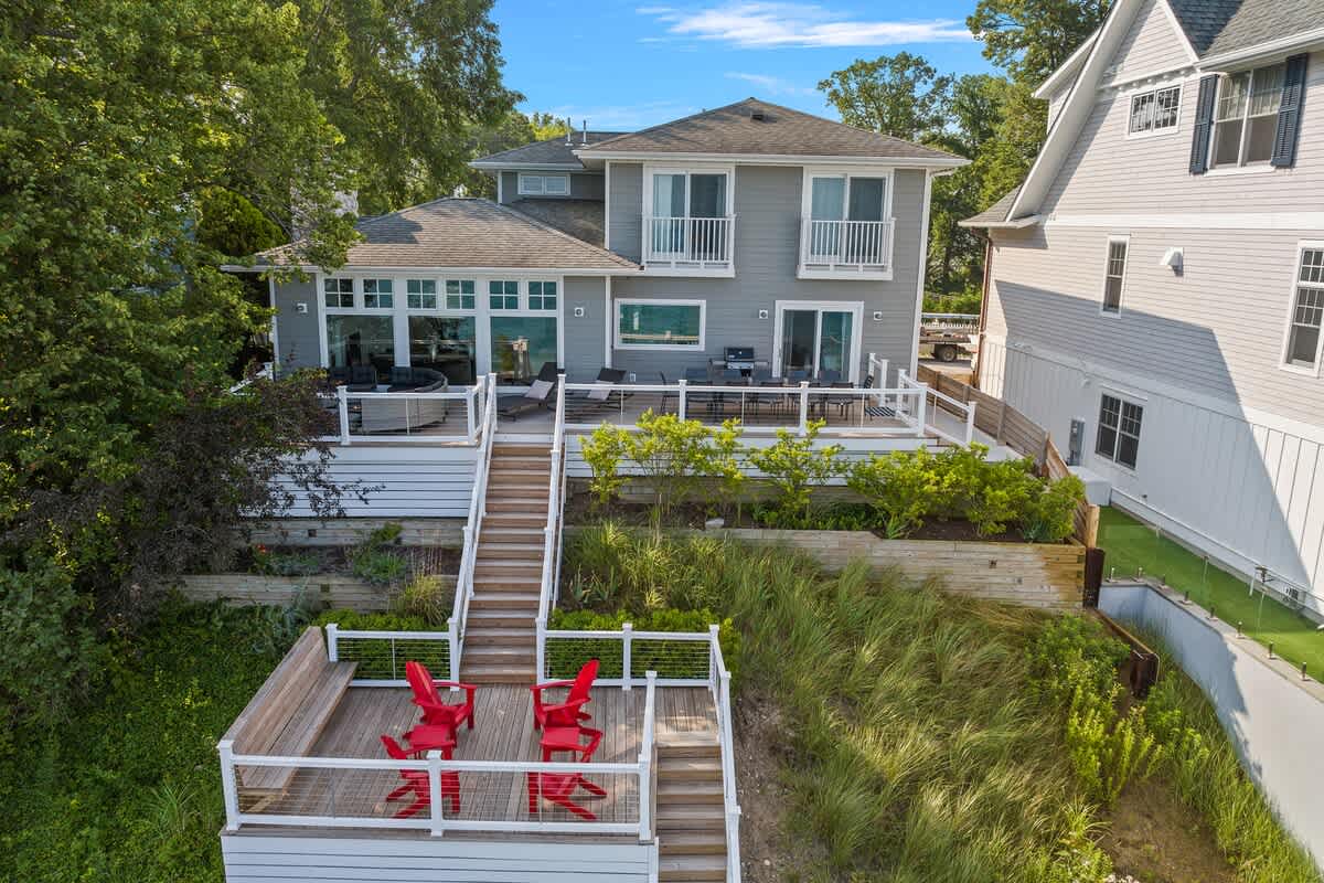 Rear exterior view of a gray house with multiple decks and stairs leading down a grassy bluff to a lower deck with red Adirondack chairs. The upper deck features outdoor seating and a grill, all overlooking the lake.