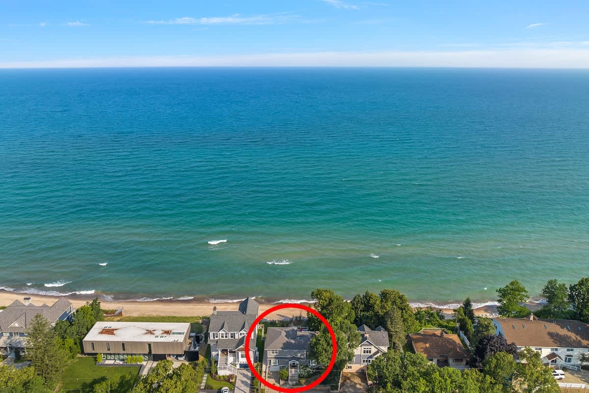 A high aerial shot captures the property, highlighted by a red circle, situated directly on the shoreline of a large blue lake. Surrounding houses and lush green trees line the coast under a clear sky.