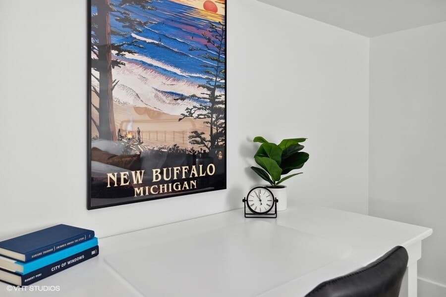 A bright white desk area features a framed "New Buffalo Michigan" poster, a small potted plant, and a vintage-style clock. A stack of books rests on the left, suggesting a quiet corner for work or reading.