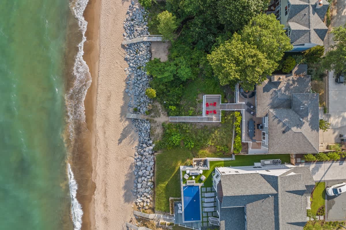 An overhead aerial view reveals the property's direct access to a sandy beach and the clear blue lake, with a long set of stairs leading down the lush, green hillside. The house and neighboring properties are nestled among dense trees.