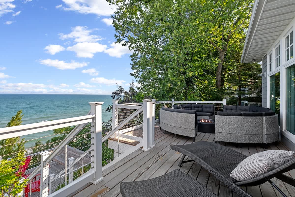 A multi-level wooden deck features a curved outdoor seating area with a fire pit, along with two lounge chairs, all overlooking the expansive blue lake. White railings with cable infill provide safety and unobstructed views.