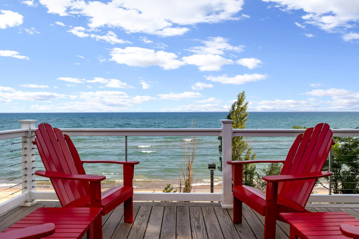 Outdoor deck with two bright red Adirondack chairs facing a vast lake with gentle waves under a blue sky with white clouds. A glass railing provides an unobstructed view of the water and distant shoreline.