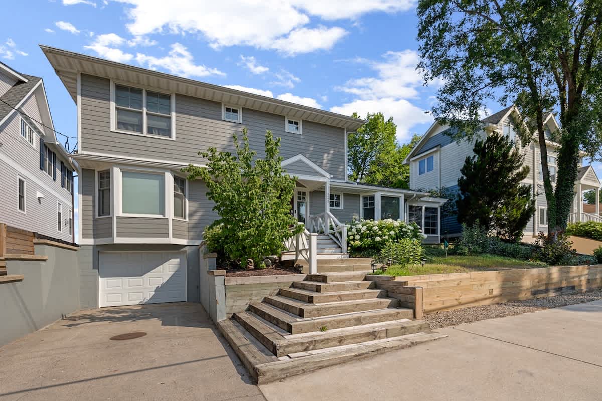 The front exterior of a grey multi-story house is visible from the street, featuring a garage, bay windows, and a prominent set of wooden steps leading to the main entrance. Lush landscaping with white flowers frames the property.