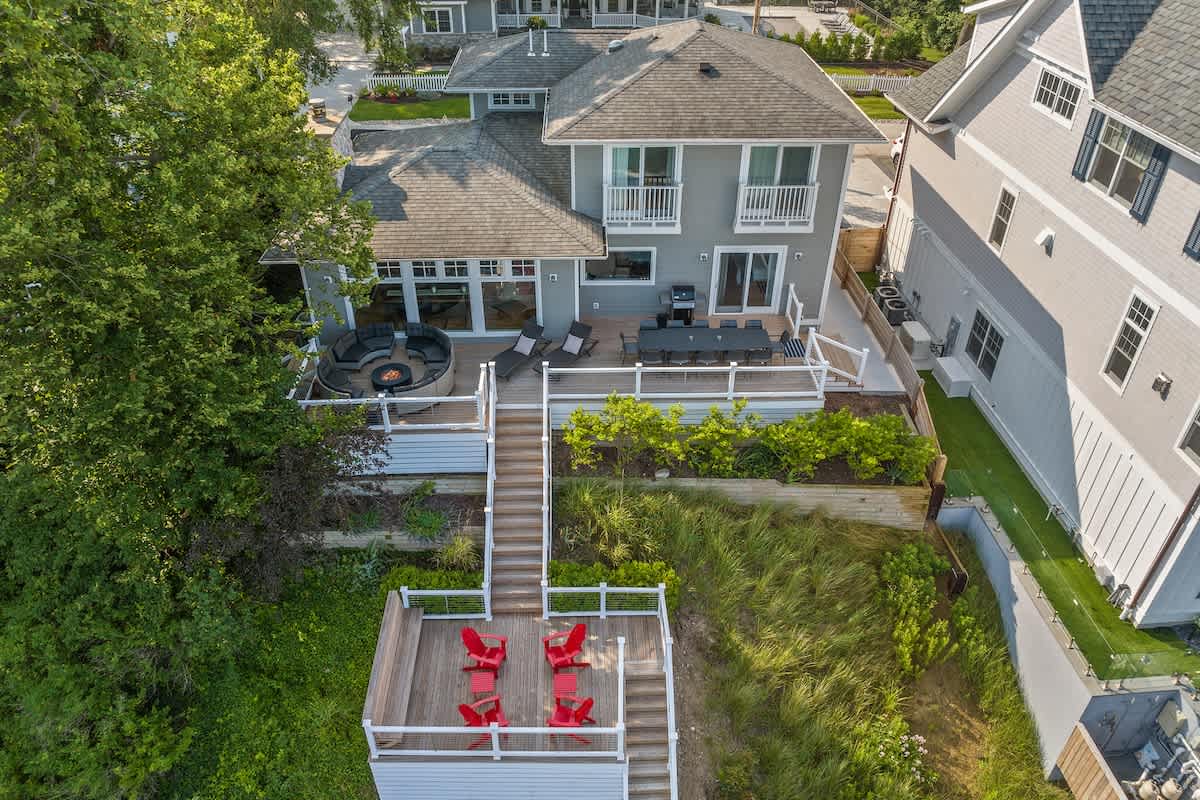 Overhead aerial view of a gray house with multiple decks and stairs descending a lush green bluff towards the lake. An outdoor fire pit seating area is visible on the main deck, and a lower deck has red chairs.