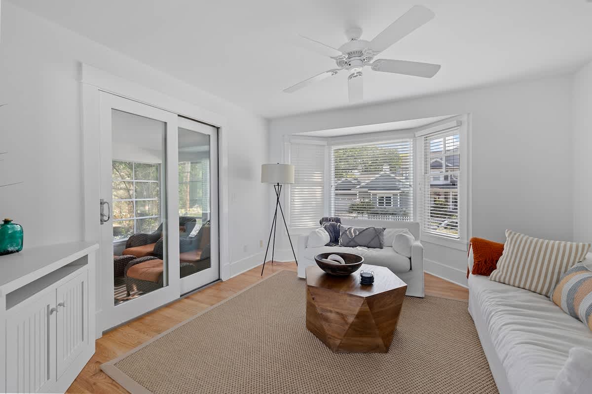 Cozy secondary living area with a white sofa, a unique hexagonal wooden coffee table, and a bay window fitted with white blinds. A sliding glass door reflects an outdoor patio, adding to the bright ambiance.