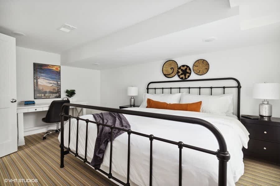 Spacious bedroom featuring a black metal bed, white bedding, black nightstands, and a white desk with a black office chair in the corner, under a framed poster.