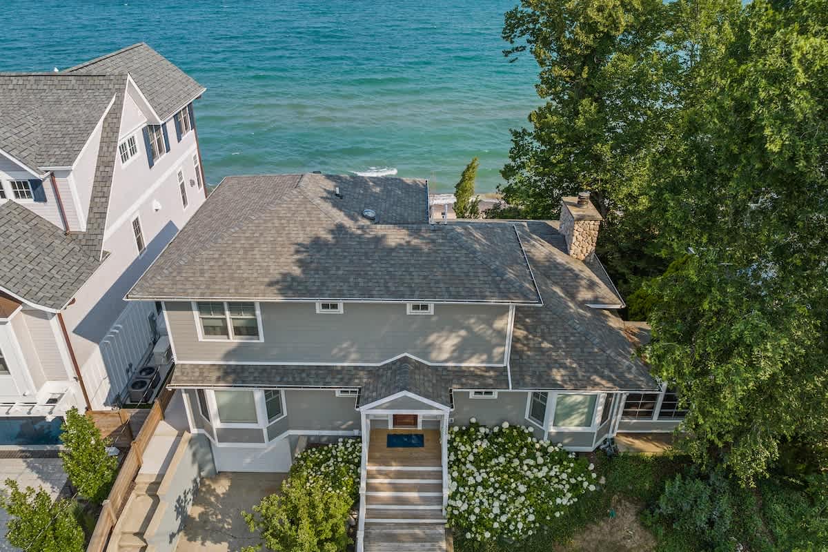 An aerial view shows the front of a grey multi-story house with a dark shingle roof, surrounded by lush green trees and white flowering bushes. The house features bay windows and a welcoming entrance with steps.