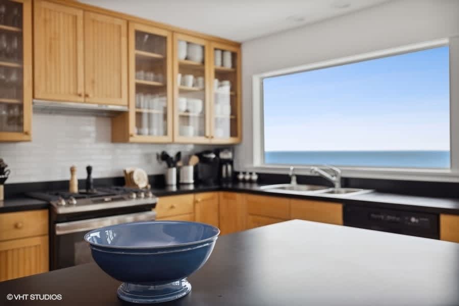 A modern kitchen features light wood cabinetry, black countertops, and stainless steel appliances, with a large window offering a direct view of the lake. A blue bowl sits on the island.