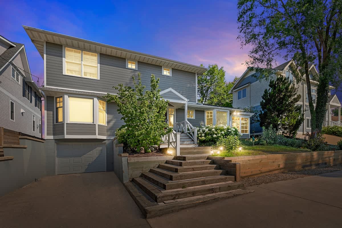 Modern gray two-story house with white trim and large windows, featuring a multi-level entrance with wooden steps and retaining walls, illuminated by warm outdoor lighting under a vibrant twilight sky.